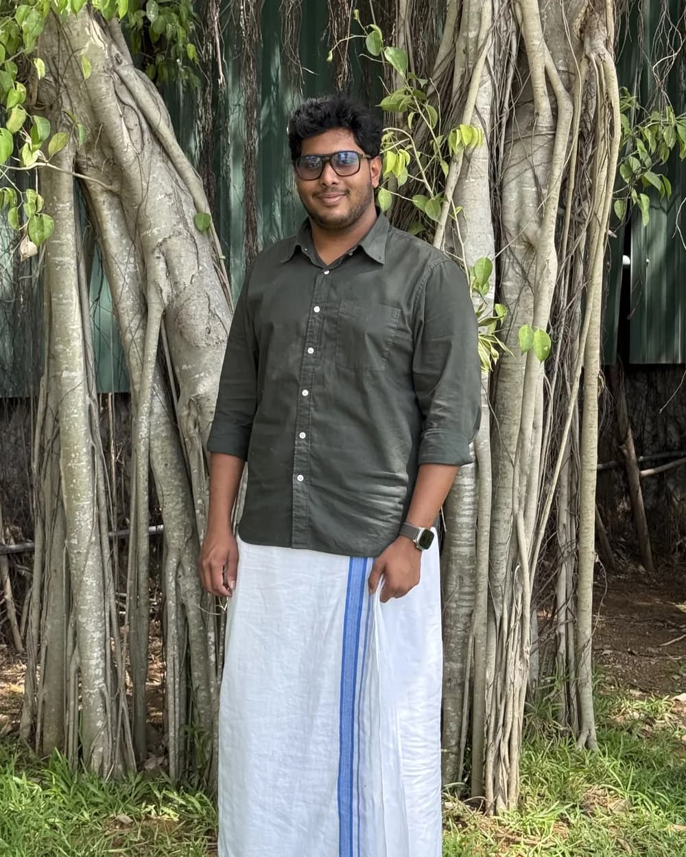 Adil in a green shirt and a white mundu, standing in front of a banyan tree